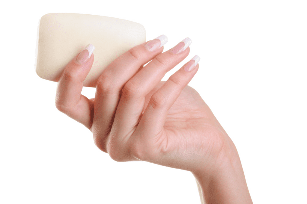 Detalhe mostrando uma mão feminina de pele clara e unhas bem feitas, segurando um sabonete em barra branco. O fundo é todo da cor branca. Imagem: Freepik.com.