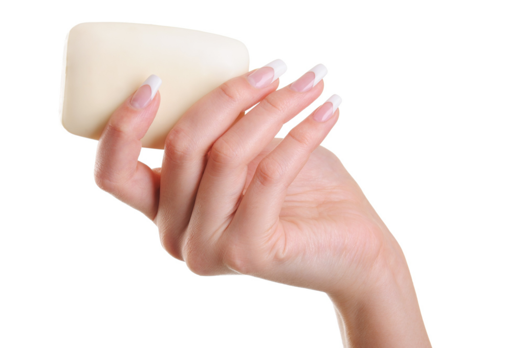 Detalhe mostrando uma mão feminina de pele clara e unhas bem feitas, segurando um sabonete em barra branco. O fundo é todo da cor branca. Imagem: Freepik.com.